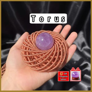 May include: A copper-colored torus-shaped orgonite with a purple amethyst sphere at its center. The word "Torus" is displayed in white block letters above the orgonite. A gift box graphic shows a purple sphere on a wooden stand.