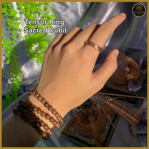 Pode incluir: Um anel Tensor cor de cobre em uma mão, com uma pulseira combinando de contas marrons. O anel é usado em um dedo. O texto "Tensor Ring Sacred Cubit" é visível. O fundo inclui estruturas decorativas de arame de cobre e vegetação.