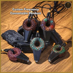 Puede incluir: Cinco colgantes conectables de frecuencia personalizada con bases negras y tapas en forma de rosquilla envueltas en alambre de colores. Los colgantes están sobre piedras de turmalina negra y una superficie de madera. El texto "Custom Frequency Connectable Pendant" está en la parte superior.