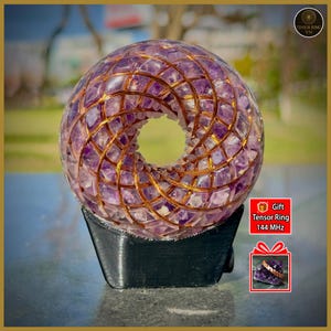 Può includere: Una scultura sferica in cristallo di ametista con un foro centrale, racchiusa in una griglia di filo di rame. La scultura poggia su una base nera. L'immagine include il testo "Tensor Ring 144 MHz" e una grafica a forma di confezione regalo.