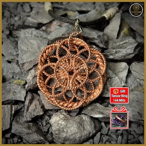 Puede incluir: Un collar con colgante de color cobre con un diseño calado circular. El colgante cuelga de un cordón oscuro con un acento beige. El collar se muestra sobre un lecho de rocas grises. La imagen incluye el texto "Tensor Ring 144 MHz".
