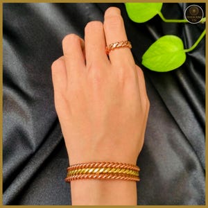 May include: A hand displays a copper-colored chain ring and a matching bracelet. The bracelet has a gold-colored band in the middle. The jewelry is set against a black background.