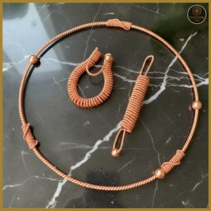 May include: A copper-colored jewelry set featuring a necklace, a ring, and a pendant. The necklace is a circular design with a twisted rope texture, accented with small copper beads and woven details. The ring and pendant have a similar twisted rope design.