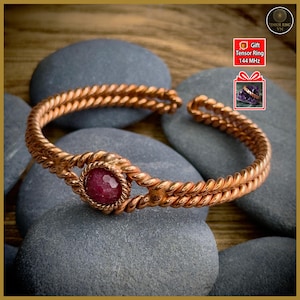 Könnte beinhalten: Kupferfarbenes Armband mit geflochtenem Seildesign. Das Armband hat einen zentralen, runden, roten Edelstein. Der Hintergrund umfasst graue Steine und den Text "Gift Tensor Ring 144 MHz".
