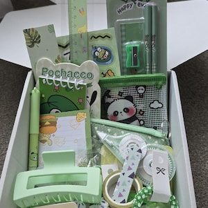 May include: A box filled with green stationery and accessories. Items include a ruler, pencils, a pencil sharpener, stickers, a hair clip, a pen, a small notebook, a coin purse, washi tape, and a bracelet. The box has a cute, kawaii aesthetic.