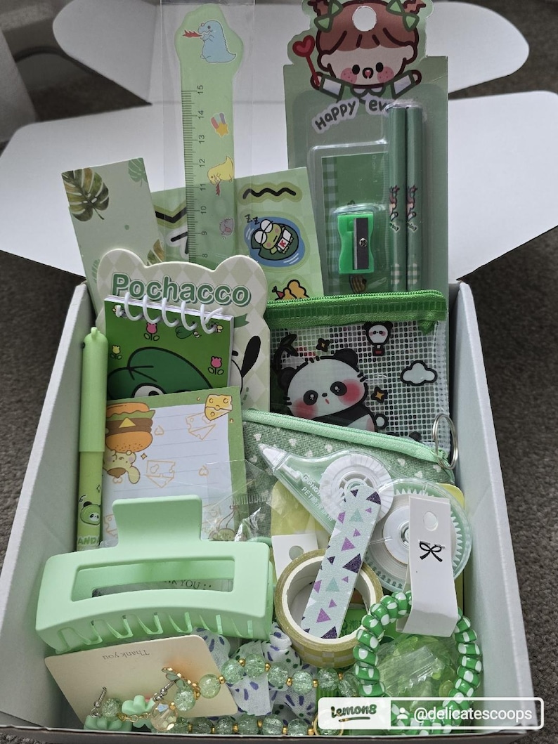 May include: A box filled with green stationery and accessories. Items include a ruler, pencils, a pencil sharpener, stickers, a hair clip, a pen, a small notebook, a coin purse, washi tape, and a bracelet. The box has a cute, kawaii aesthetic.