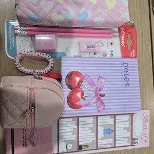 May include: A collection of pink stationery items, including a pencil case with heart patterns, pencils, an eraser, a hair tie, a small purse, a notebook with cherry illustrations, and a bookmark.