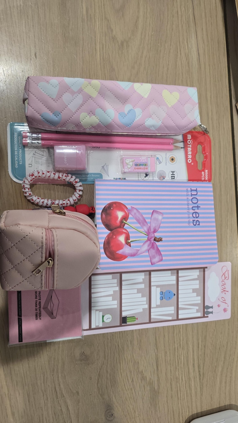 May include: A collection of pink stationery items, including a pencil case with heart patterns, pencils, an eraser, a hair tie, a small purse, a notebook with cherry illustrations, and a bookmark.