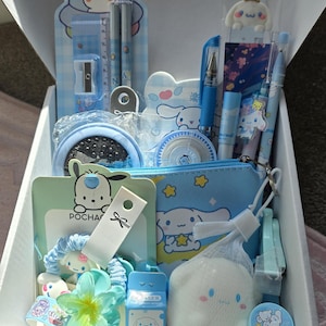 May include: A white box filled with blue and white stationery and accessories. Items include pens, a pencil sharpener, a ruler, a small pouch, a hair scrunchie, a small eraser, and a decorative sponge. The box is open, revealing the contents.