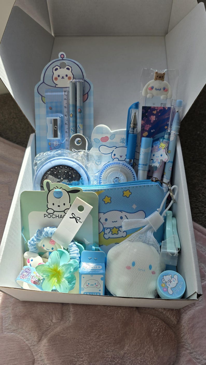May include: A white box filled with blue and white stationery and accessories. Items include pens, a pencil sharpener, a ruler, a small pouch, a hair scrunchie, a small eraser, and a decorative sponge. The box is open, revealing the contents.