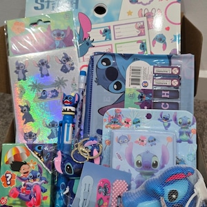 May include: A box filled with various Stitch-themed school supplies and accessories. Items include notebooks, stickers, a pen, a pencil case, and a plush toy. The color scheme is primarily blue, pink, and white, featuring the character Stitch from Disney's Lilo & Stitch.