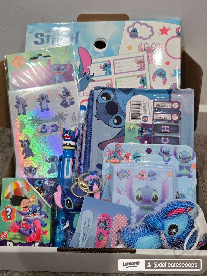 May include: A box filled with various Stitch-themed school supplies and accessories. Items include notebooks, stickers, a pen, a pencil case, and a plush toy. The color scheme is primarily blue, pink, and white, featuring the character Stitch from Disney's Lilo & Stitch.
