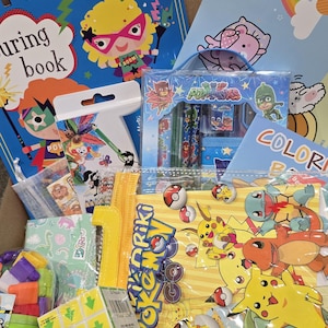 May include: A box filled with children's books, coloring books, and stationery. The items include a superhero-themed book, a coloring book with a rainbow, and a set of PJ Masks pencils. There are also Pokemon-themed stickers and a Rubik's cube.