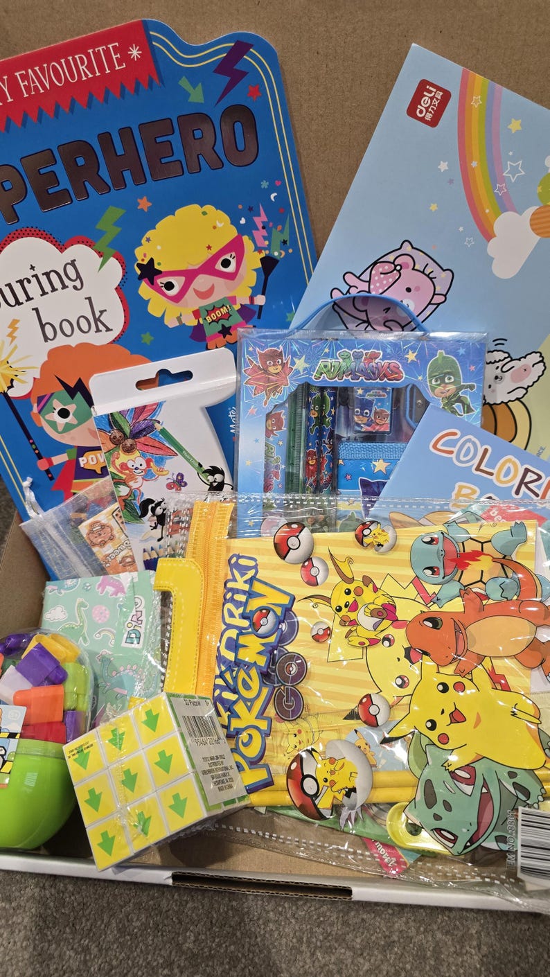 May include: A box filled with children's books, coloring books, and stationery. The items include a superhero-themed book, a coloring book with a rainbow, and a set of PJ Masks pencils. There are also Pokemon-themed stickers and a Rubik's cube.