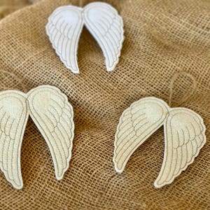 May include: Three decorative angel wing ornaments. The wings are off-white with gold stitching and a jute string for hanging. They are arranged on a textured, tan burlap background.