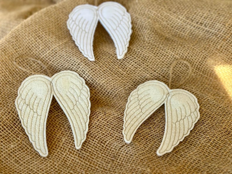 May include: Three decorative angel wing ornaments. The wings are off-white with gold stitching and a jute string for hanging. They are arranged on a textured, tan burlap background.