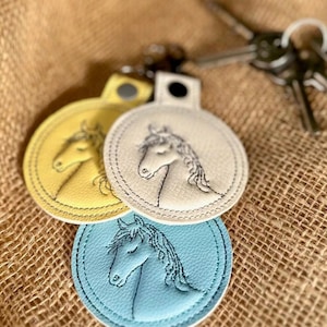 May include: Three round keychains in yellow, cream, and light blue, each featuring an embroidered horse head design. The keychains are made of leather-like material and are on a textured surface. Keys are visible in the background.