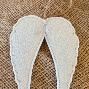 May include: A pair of off-white felt angel wings with a stitched outline and a jute string for hanging. The wings have a scalloped edge and are displayed against a textured, brown background.