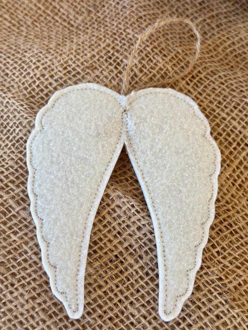 May include: A pair of off-white felt angel wings with a stitched outline and a jute string for hanging. The wings have a scalloped edge and are displayed against a textured, brown background.