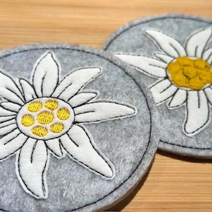 Puede incluir: Dos posavasos redondos de fieltro gris con diseños bordados de flores de edelweiss blancas y amarillas. Los posavasos tienen un borde negro cosido y están sobre una superficie de madera.