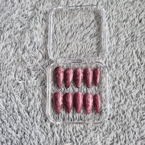 May include: A set of ten burgundy glitter press-on nails in a clear plastic case. Each nail has a pointed shape and a floral design. The nails are arranged in two rows of five. The nails are a deep red colour.