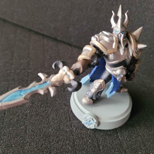 May include: A painted miniature figurine of a warrior wearing silver armor with gold accents, a blue cape, and a horned helmet. The figure holds a long sword with a skull-shaped hilt and a glowing blue blade, standing on a circular gray base.