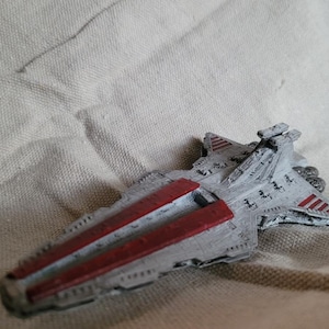 May include: A miniature starship model featuring a wedge-shaped hull painted in light grey with prominent dark red panels along the top deck. The spacecraft includes a raised command bridge, detailed surface panelling, and rear engine thrusters.