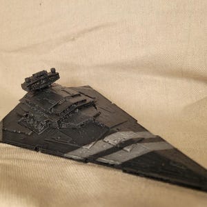 May include: A black and grey model of a Star Destroyer spaceship. The model has detailed features, including turrets and layered sections. Grey stripes accent the ship's angled hull. The model is set against a neutral fabric backdrop.