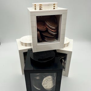 May include: A collection of three square coin banks constructed from building blocks. Two white banks are positioned behind one black bank. Each bank features a clear front panel revealing various metal coins stored inside, including copper and silver.