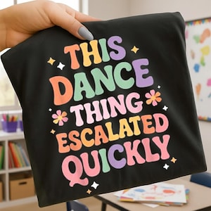 Puede incluir: Camiseta negra con la frase "THIS DANCE THING ESCALATED QUICKLY" en letras coloridas de estilo retro. El diseño incluye estrellas y flores. La camiseta está sujeta por una mano.