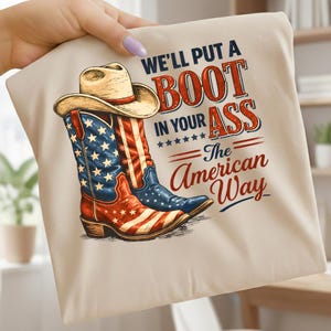 May include: A beige t-shirt featuring a graphic of cowboy boots in red, white, and blue, with a cowboy hat above. The text reads "WE'LL PUT A BOOT IN YOUR ASS - The American Way."