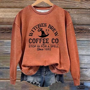 Può includere: Una felpa a girocollo a maniche lunghe color ruggine con testo nero che recita Witches Brew Coffee Co, Stop In For A Spell, Since 1692, e un grafico di un cappello da strega con stelle. Il capo è esposto su una gruccia sopra un paio di pantaloncini di jeans.