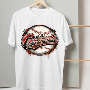 May include: A white short-sleeved t-shirt featuring a graphic of a baseball with red stitching. The word Cardinals is written in a stylized red and black font across the center, with a silhouette of an arch visible above the text.