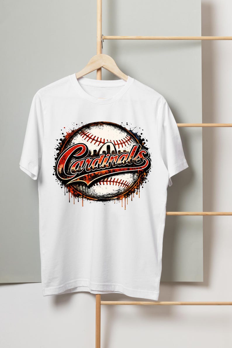 May include: A white short-sleeved t-shirt featuring a graphic of a baseball with red stitching. The word Cardinals is written in a stylized red and black font across the center, with a silhouette of an arch visible above the text.