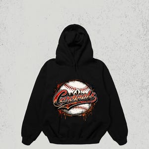 May include: A black hooded sweatshirt featuring a graphic print of a baseball with red stitching. The word Cardinals is written in a stylized red and white font across the center of the baseball, with orange and black paint splatter effects.
