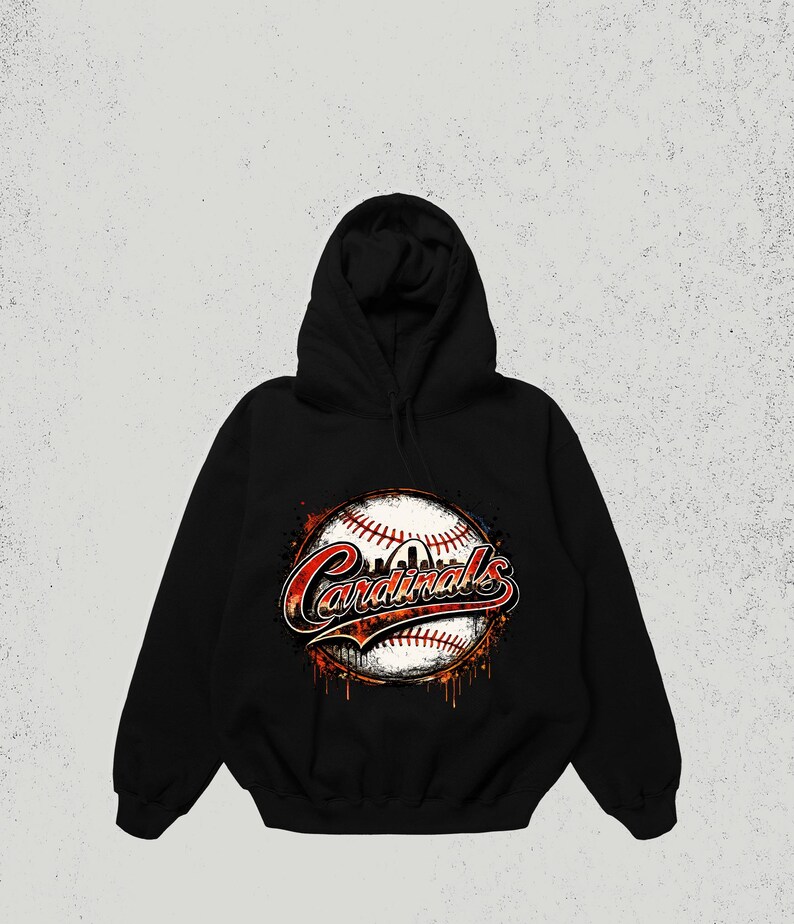 May include: A black hooded sweatshirt featuring a graphic print of a baseball with red stitching. The word Cardinals is written in a stylized red and white font across the center of the baseball, with orange and black paint splatter effects.