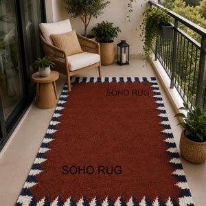 Pu&ograve; includere: Un tappeto rettangolare con un centro color ruggine e un bordo di triangoli bianchi e blu navy alternati. Il testo "SOHO RUG" &egrave; stampato sul tappeto. Il tappeto &egrave; su un balcone con una sedia, piante e un tavolino.