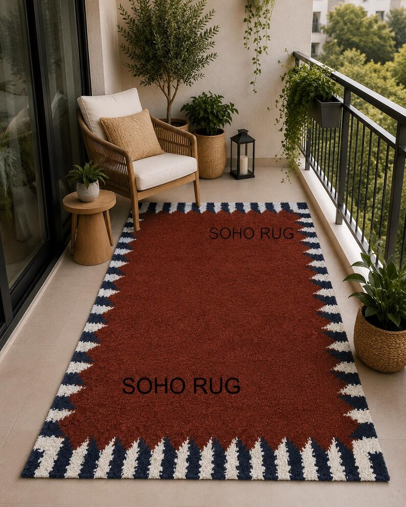 Pu&ograve; includere: Un tappeto rettangolare con un centro color ruggine e un bordo di triangoli bianchi e blu navy alternati. Il testo "SOHO RUG" &egrave; stampato sul tappeto. Il tappeto &egrave; su un balcone con una sedia, piante e un tavolino.