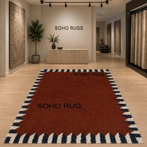 Pu&ograve; includere: Un tappeto rettangolare color ruggine con un bordo a zig-zag bianco e blu navy. Il tappeto &egrave; esposto in uno showroom con il testo "SOHO RUGS" visibile sulla parete. Il tappeto &egrave; il punto focale, che mostra un design moderno.