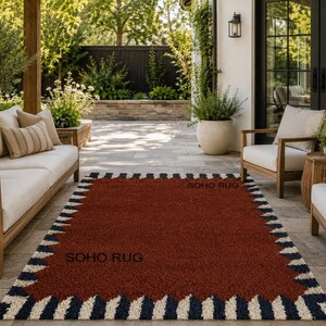 Pu&ograve; includere: Un tappeto rettangolare con un centro rosso intenso e un bordo a motivi di zigzag blu navy e crema alternati. Il testo "SOHO RUG" &egrave; visibile sul tappeto. Il tappeto &egrave; posizionato su un patio con mobili da esterno.