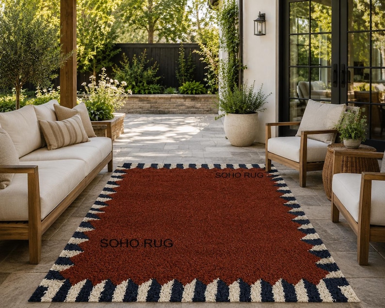 Pu&ograve; includere: Un tappeto rettangolare con un centro rosso intenso e un bordo a motivi di zigzag blu navy e crema alternati. Il testo "SOHO RUG" &egrave; visibile sul tappeto. Il tappeto &egrave; posizionato su un patio con mobili da esterno.