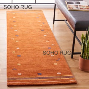 May include: An orange rectangular rug with a textured surface. The rug features small, scattered geometric patterns in white, blue, and yellow. The rug has a border with blue and yellow stripes. The text "SOHO RUG" is visible.