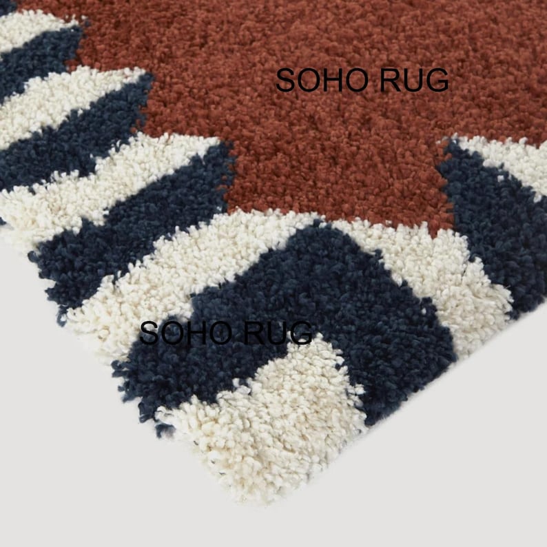 Pu&ograve; includere: Primo piano di un tappeto strutturato con un motivo geometrico. Il tappeto presenta una sezione centrale di colore marrone rossiccio, con sezioni a righe bianche e blu navy. Il testo "SOHO RUG" &egrave; visibile sul tappeto.