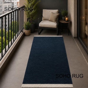 Op de afbeelding: Een marineblauw rechthoekig tapijt met een crèmekleurige franje ligt op een balkon. Het tapijt is gemaakt van een textuurrijk materiaal. De woorden "SOHO RUG" staan op het tapijt gedrukt. Een stoel, een kussen en een kleine tafel zijn ook zichtbaar.