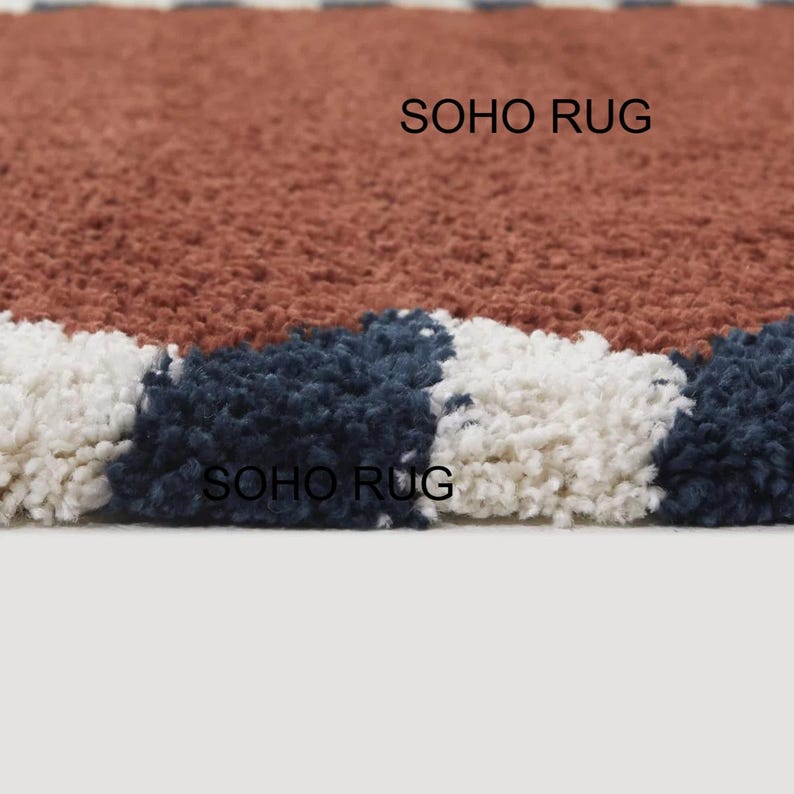 Pu&ograve; includere: Primo piano di un tappeto strutturato con un design a blocchi di colore. Il tappeto presenta una combinazione di ruggine, blu navy e bianco. Il testo "SOHO RUG" &egrave; stampato sul tappeto.