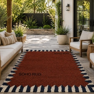 Pu&ograve; includere: Un tappeto rettangolare con un centro rosso intenso e un bordo a motivi di zigzag blu navy e crema alternati. Il testo "SOHO RUG" &egrave; visibile sul tappeto. Il tappeto &egrave; posizionato su un patio con mobili da esterno.