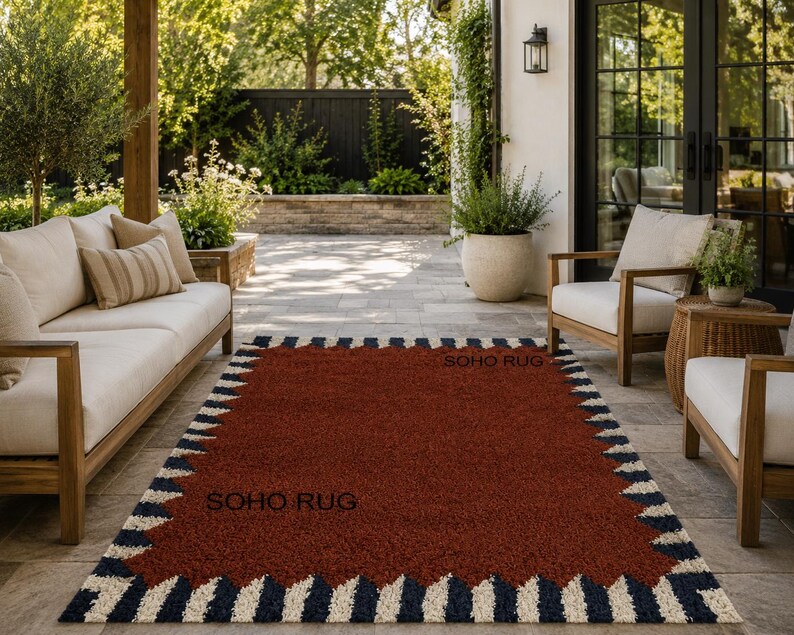 Pu&ograve; includere: Un tappeto rettangolare con un centro rosso intenso e un bordo a motivi di zigzag blu navy e crema alternati. Il testo "SOHO RUG" &egrave; visibile sul tappeto. Il tappeto &egrave; posizionato su un patio con mobili da esterno.
