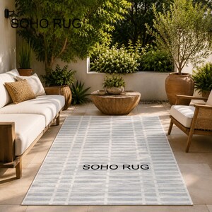 May include: A rectangular outdoor rug with a light gray and white striped pattern. The rug is on a patio with outdoor furniture and greenery. The text "SOHO RUG" is printed on the rug.