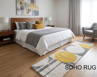 Può includere: Un tappeto rettangolare con un design geometrico in una camera da letto. Il tappeto presenta grandi cerchi gialli e bianchi, insieme a forme lineari grigie, su uno sfondo color crema. Il tappeto è posto davanti a un letto con testiera in legno.