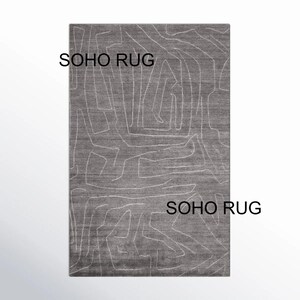 May include: A rectangular gray rug with a white abstract design. The rug has a textured surface and the text "SOHO RUG" is printed on the top and bottom of the rug.